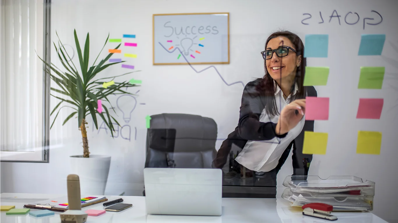 Smiling businesswoman organizing sticky notes on a glass wall in a modern office, brainstorming recruitment strategy.