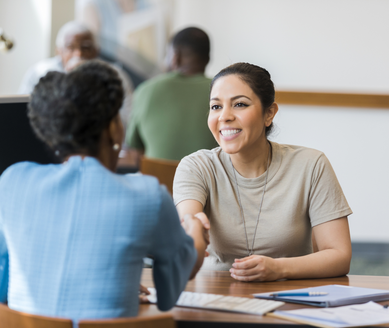Smiling woman in military-style T-shirt shaking hands with another woman in a professional setting.